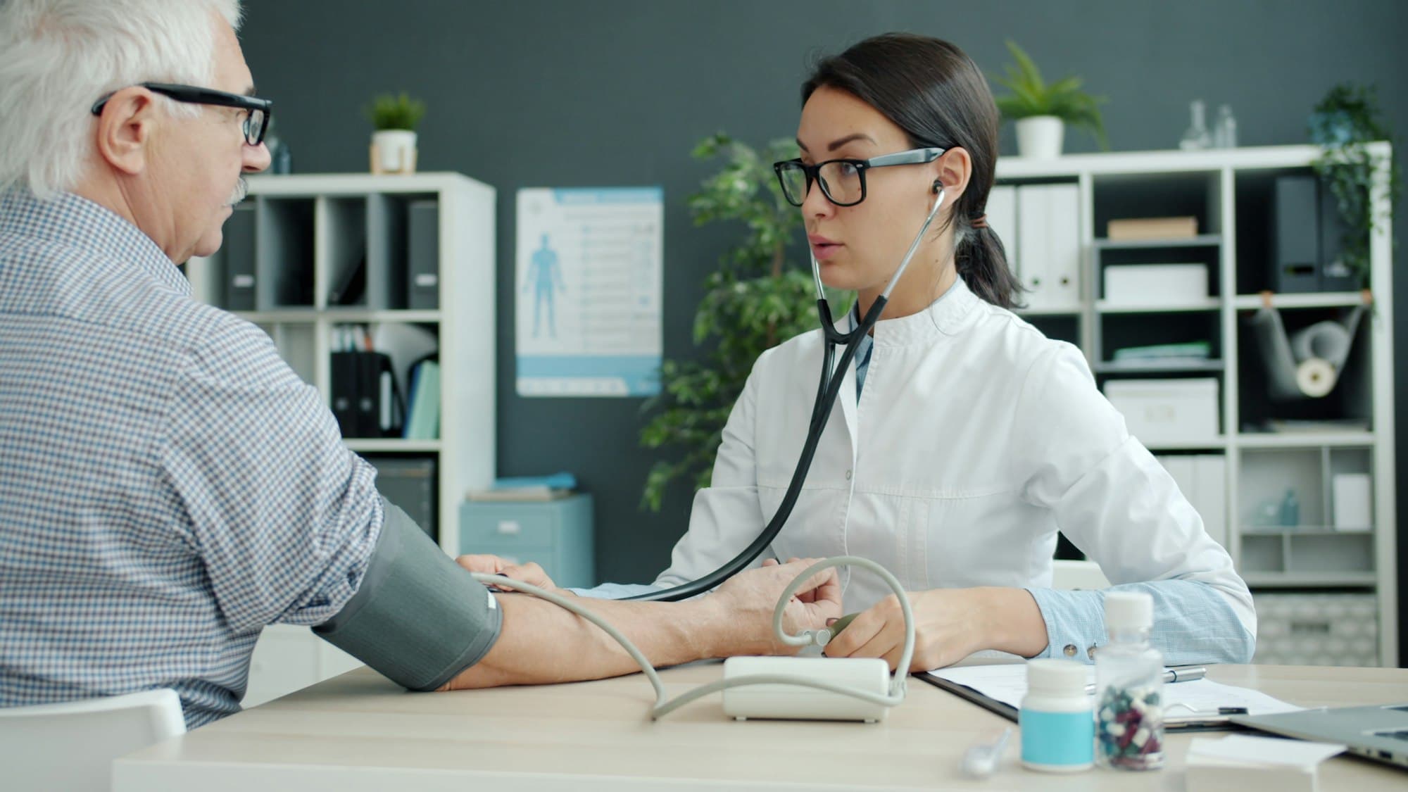 A physician checks an older patient's blood pressure across a desk in a small office