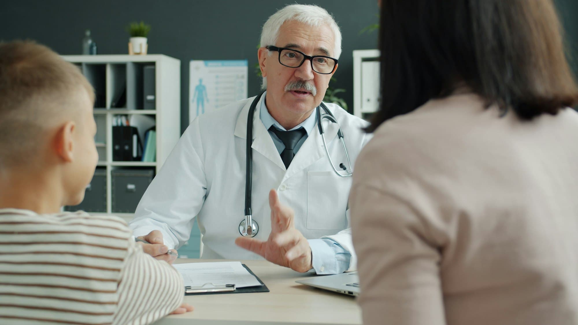 A physician talks with a parent and child across a desk in a small office