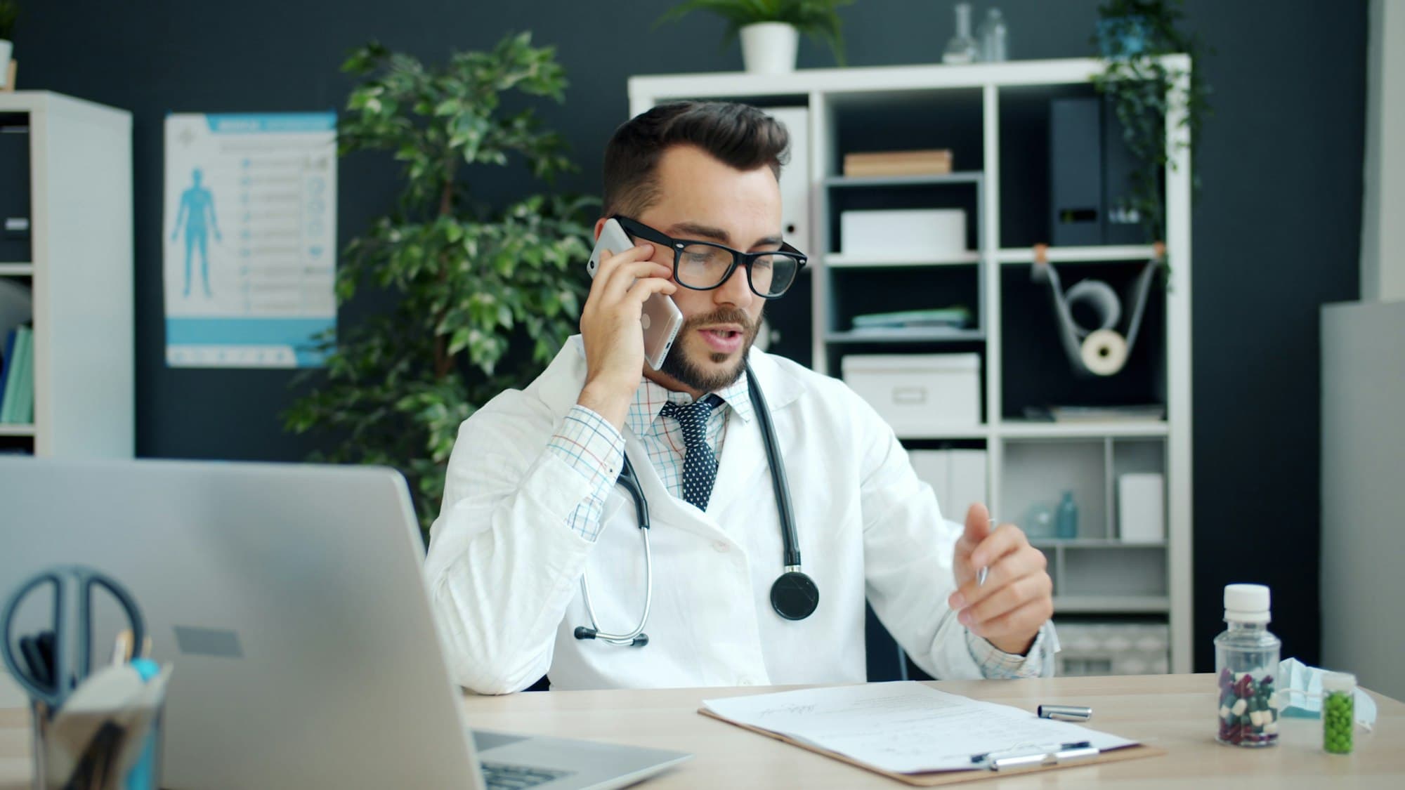 A physician sits at a desk talking on a mobile phone beside an open laptop