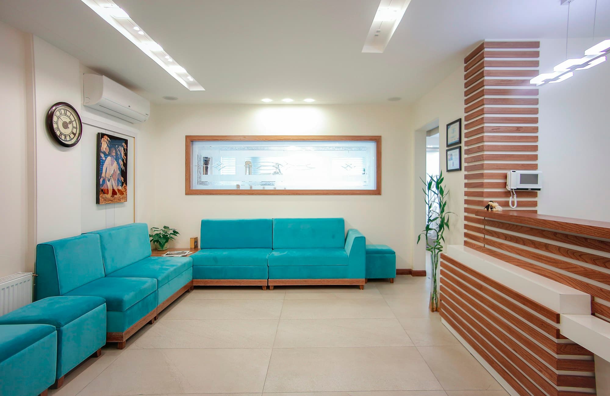 An empty clinic waiting room with teal sofas, a wall clock, and a reception desk