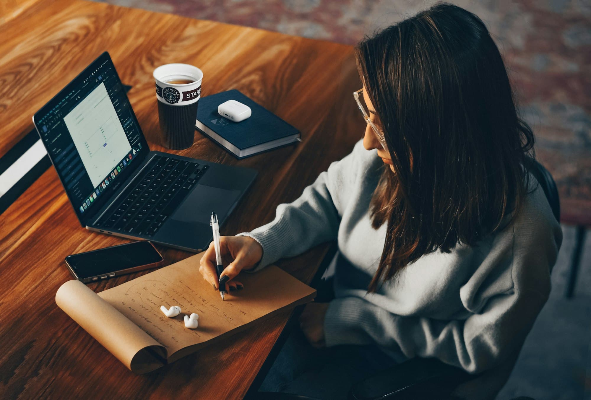 A woman writes notes by hand at a wooden desk beside an open laptop and a cup of coffee